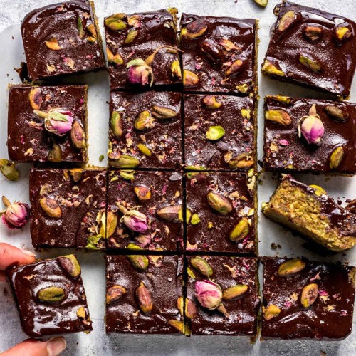 Dark Chocolate Pistachio Bars topped with pistachios and rose petals, cut into squares, with 2 fingers grabbing a piece.