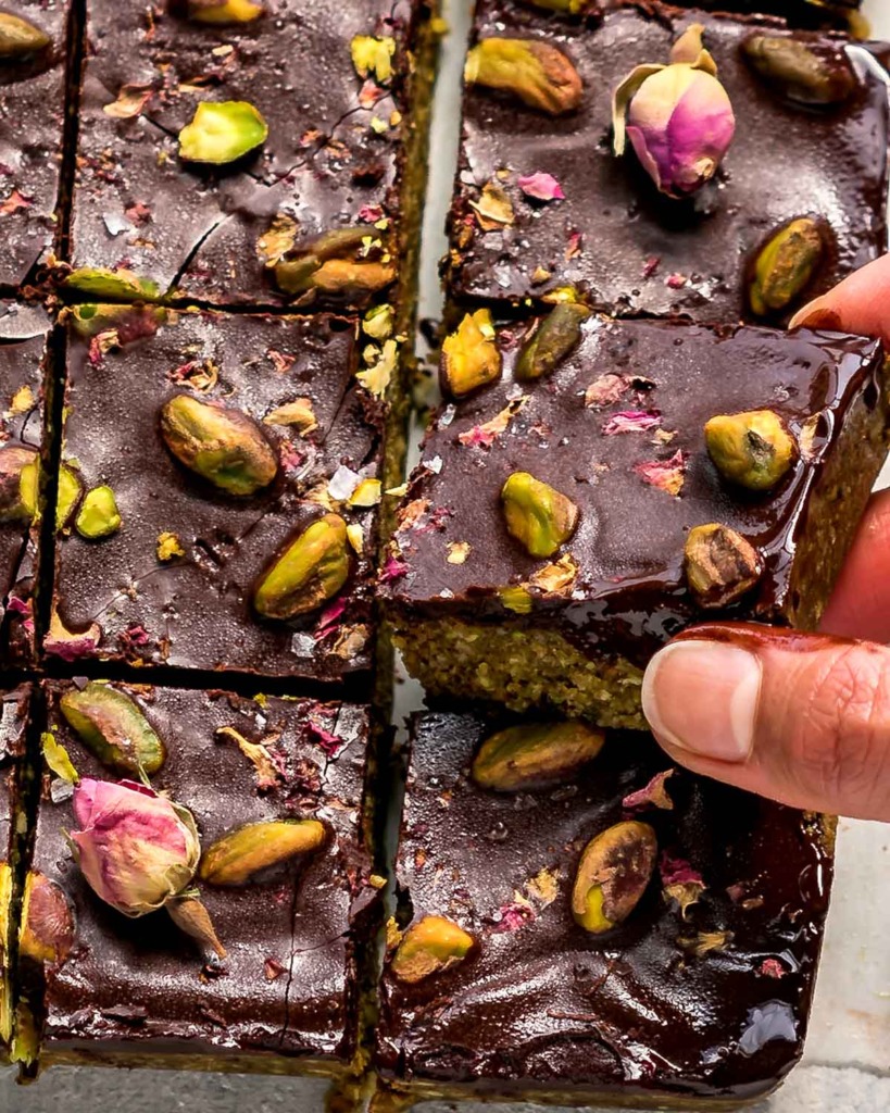 Closeup of a hand grabbing slice of Dark Chocolate Pistachio Bars topped with pistachios and rose petals