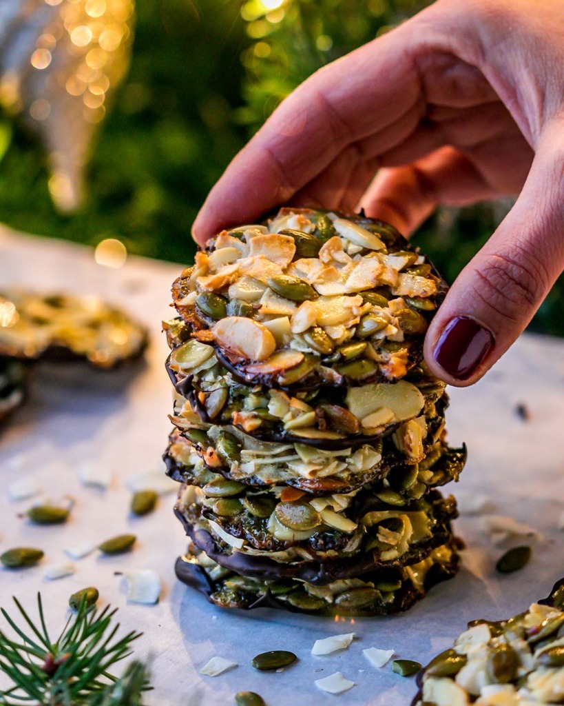 Crunchy Almond Seed Chocolate Cookies Stacked on a parchment paper lined cooling rack with nuts and seeds around and a lit up Christmas tree in the background and fingers reaching to grab the top cookie