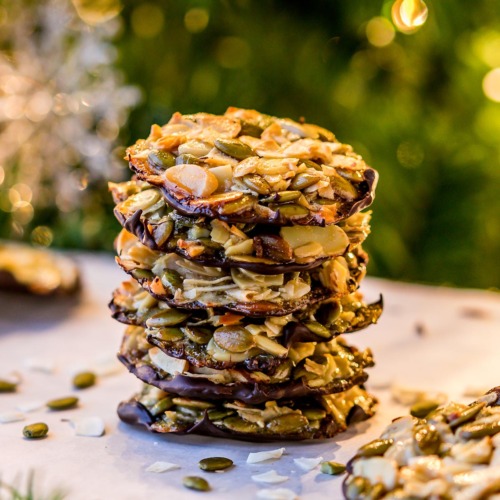Crunchy Almond Seed Chocolate Cookies Stacked on a parchment paper lined cooling rack with nuts and seeds around and a lit up Christmas tree in the background